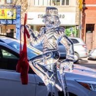 Transparent dancer ice sculpture beside a parked car on a city street, with red toe shoes draped and hanging down.