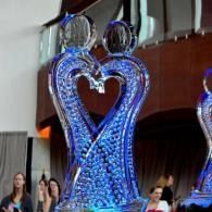 Blue glass sculpture of two figures forming a heart, displayed indoors with people in the background