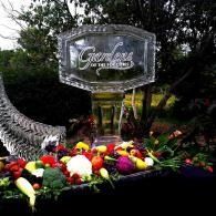 Floral memorial tribute by a clear glass sign, with red, yellow, and white flowers in a garden setting