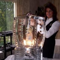 Person standing behind a lit ice sculpted display wine ice luge indoors, smiling at the camera.