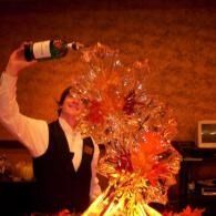 Bartender pours liquor over a flaming cocktail ice luge, creating a dramatic fireburst in a dim bar.