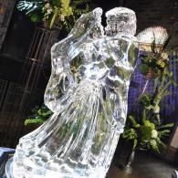 Clear ice sculpture of two figures embracing outdoors beside greenery and a building