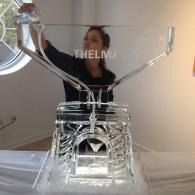 Clear ice luge sculpted giant serving glass   with a person standing behind it and the word “THELMU” across the center.