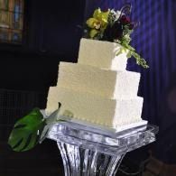 White tiered cake on a clear stand, topped with flowers and greenery in a dimly lit setting