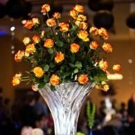 Orange roses arranged in a tall white vase at a dimly lit event