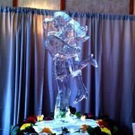 Transparent ice sculpture of a person standing on a pedestal with flowers, against draped curtains