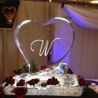 Heart-shaped ice sculpture with a scripted “W,” surrounded by red roses and draped fabric at an event display