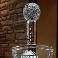 Crystal trophy with a spherical top and “CASANDRA” on the base, displayed against a brick wall.