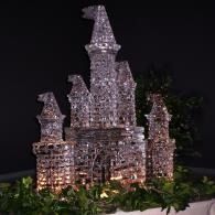 A sparkling castle made of ice with lights against a dark background, with greenery at the base