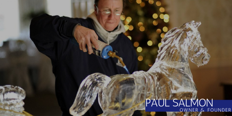 Artist carving an ice horse with a torch in a workshop, with a lit Christmas tree in the background