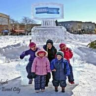 Children posing in winter coats before an ice sculpture in a snowy town square