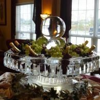 Fruit display on a dining table with grapes and decorative glassware bottles with an ice sculpture in the center by a window