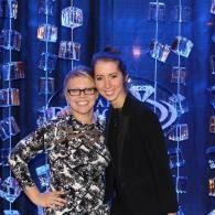 Two women posing together in front of a blue event backdrop with hanging glass decorations