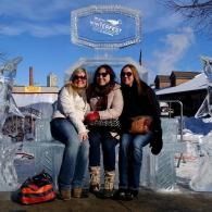 Three people seated on an icy bench outdoors on a sunny winter day, smiling and wearing sunglasses.