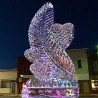 Abstract translucent winged sculpture glowing purple and pink at night in an outdoor plaza