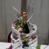 Champagne bottle in a clear ice sculpture, decorated with flowers on a table.