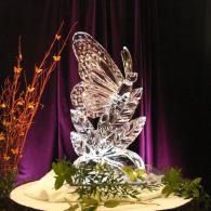 Clear crystal butterfly sculpture on a lit table with purple drapes and greenery