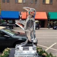 Ice sculpture on a sidewalk in front of colorful storefronts, shaped like a penguin serving a cocktail.
