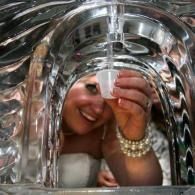 Person smiling behind a reflective metallic tunnel, holding a clear cup under running water.