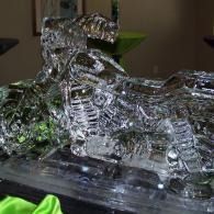 Transparent glass sculpture of a motorcycle on a dark base, with a green object in the corner