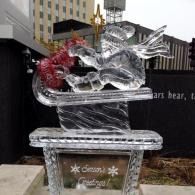 Clear ice sculpture of a child on a sled outdoors, decorated with red accents and a gold crown