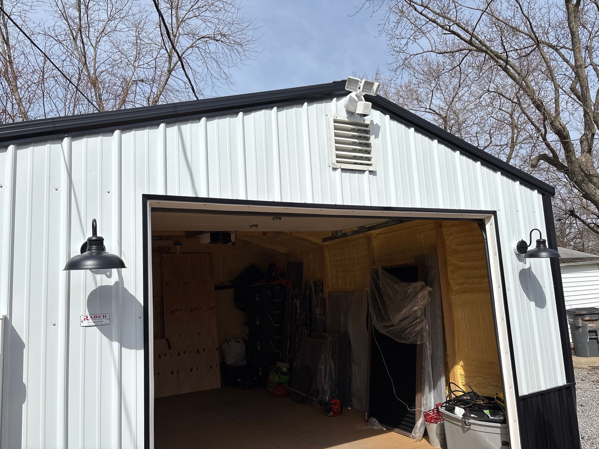 White metal garage with open door, lit inside, under leafless trees on a sunny day