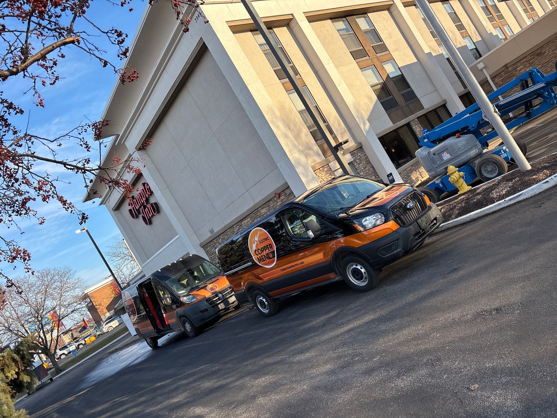 Orange-and-black truck parked beside a large building on a sunny street, with a red pickup behind it