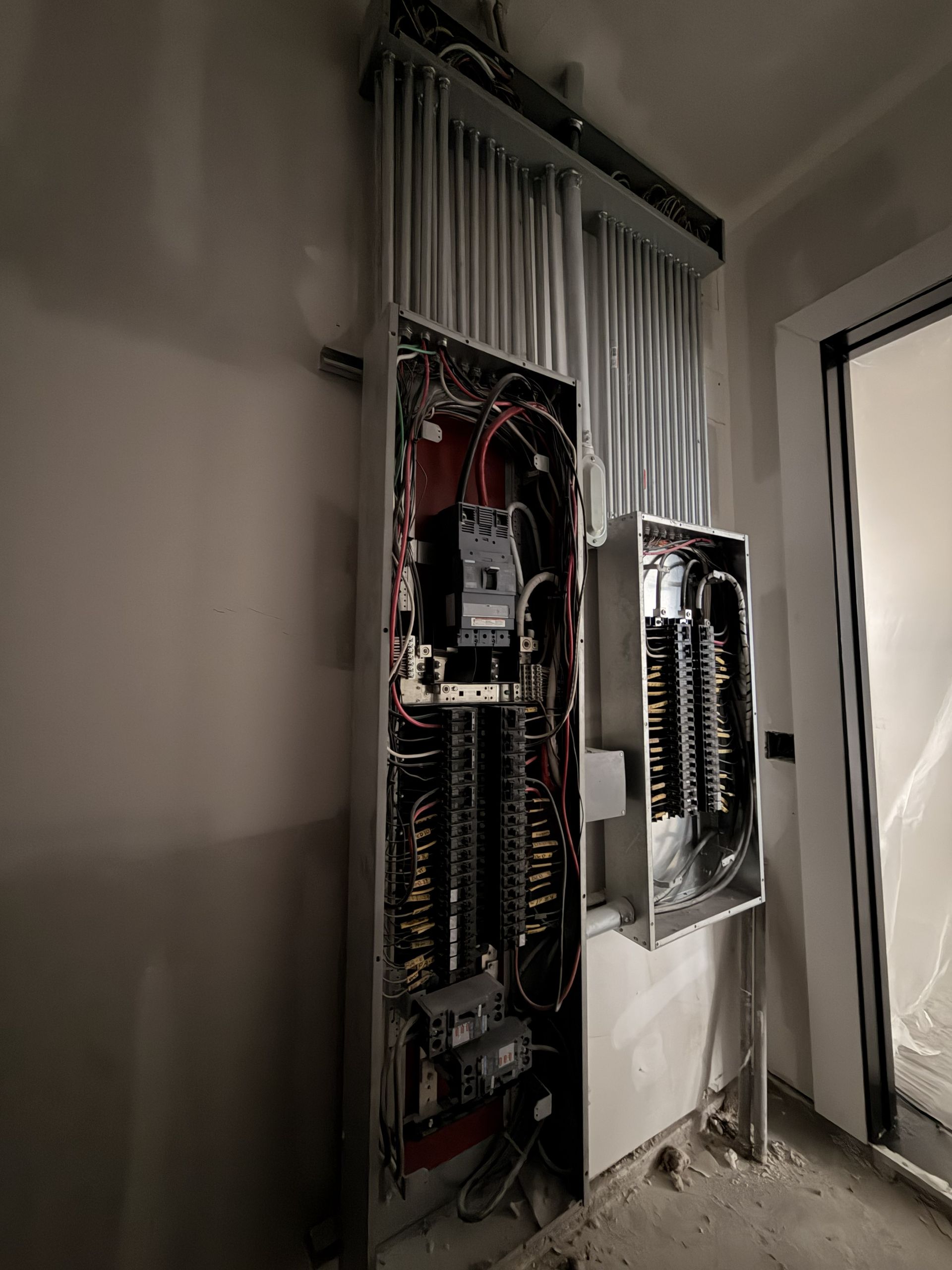 Electrical panel and exposed circuit breakers mounted on a wall in a dim utility room.