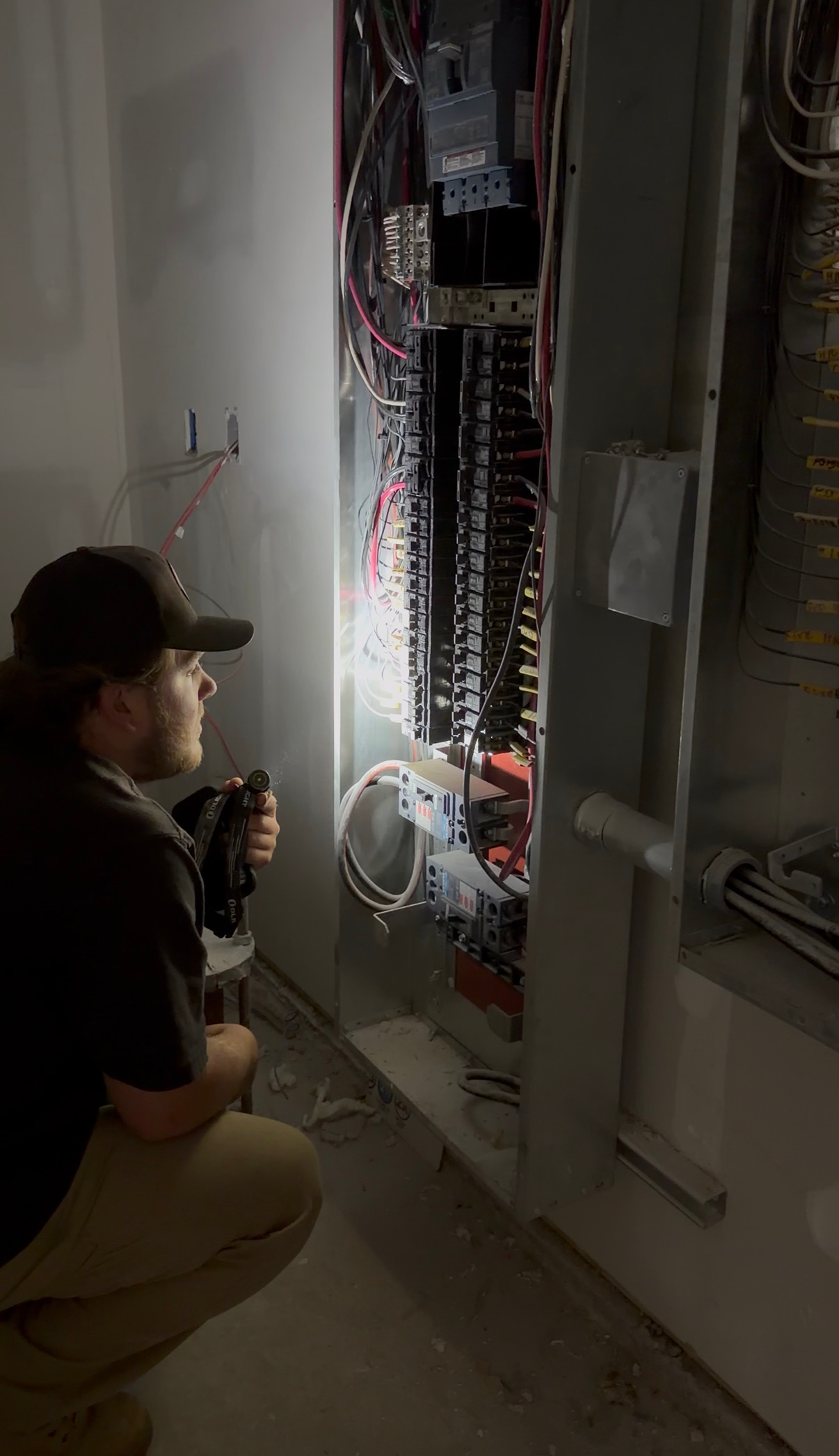 Technician kneeling beside an open electrical panel in a narrow utility corridor, inspecting wiring.