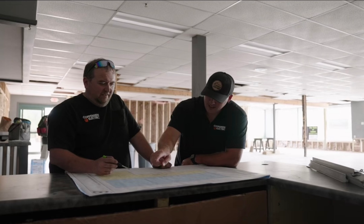 Two men in dark shirts lean over blueprints on a table in a bright workshop.