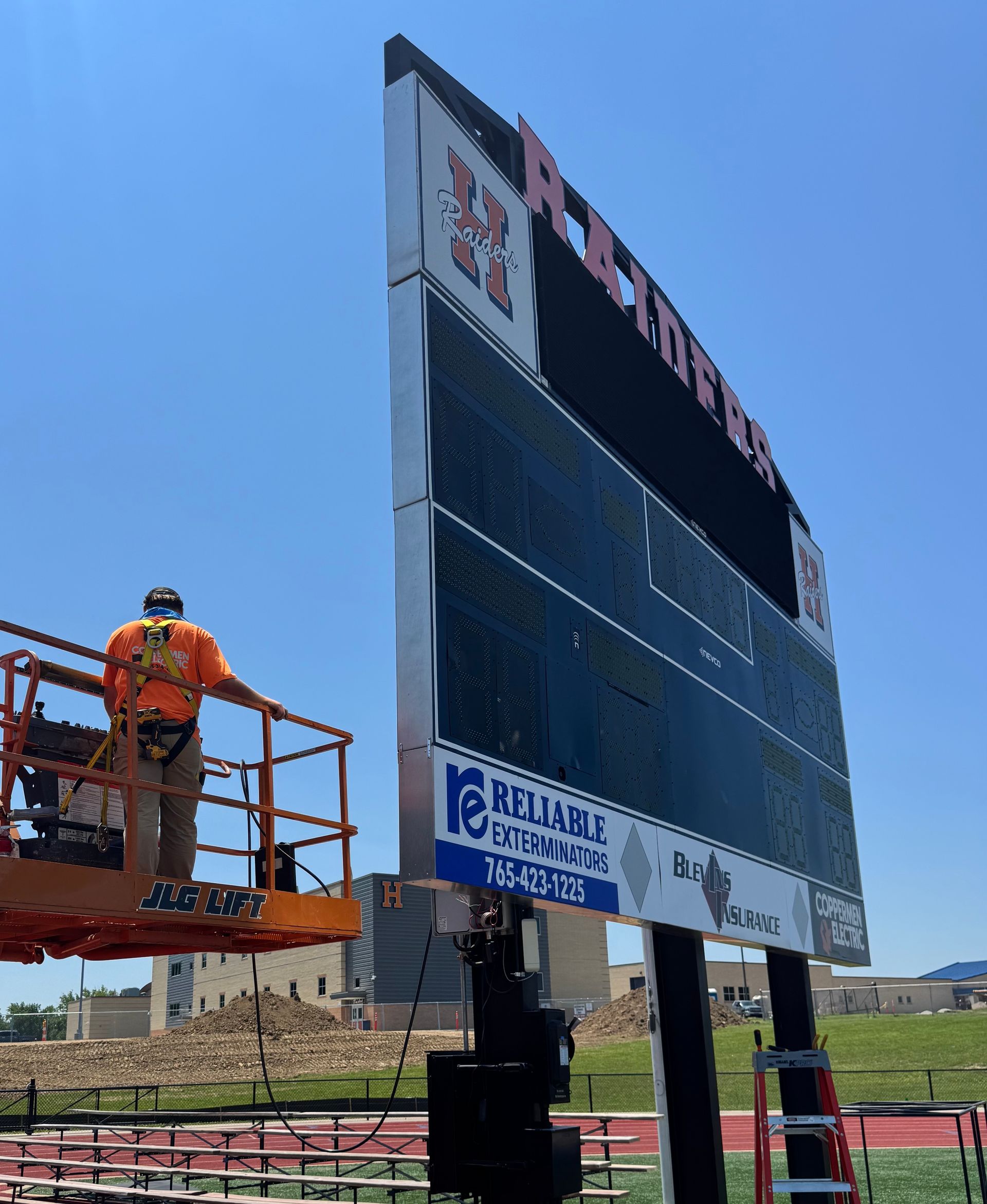 Worker on lift installing a large scoreboard outdoors on a clear day