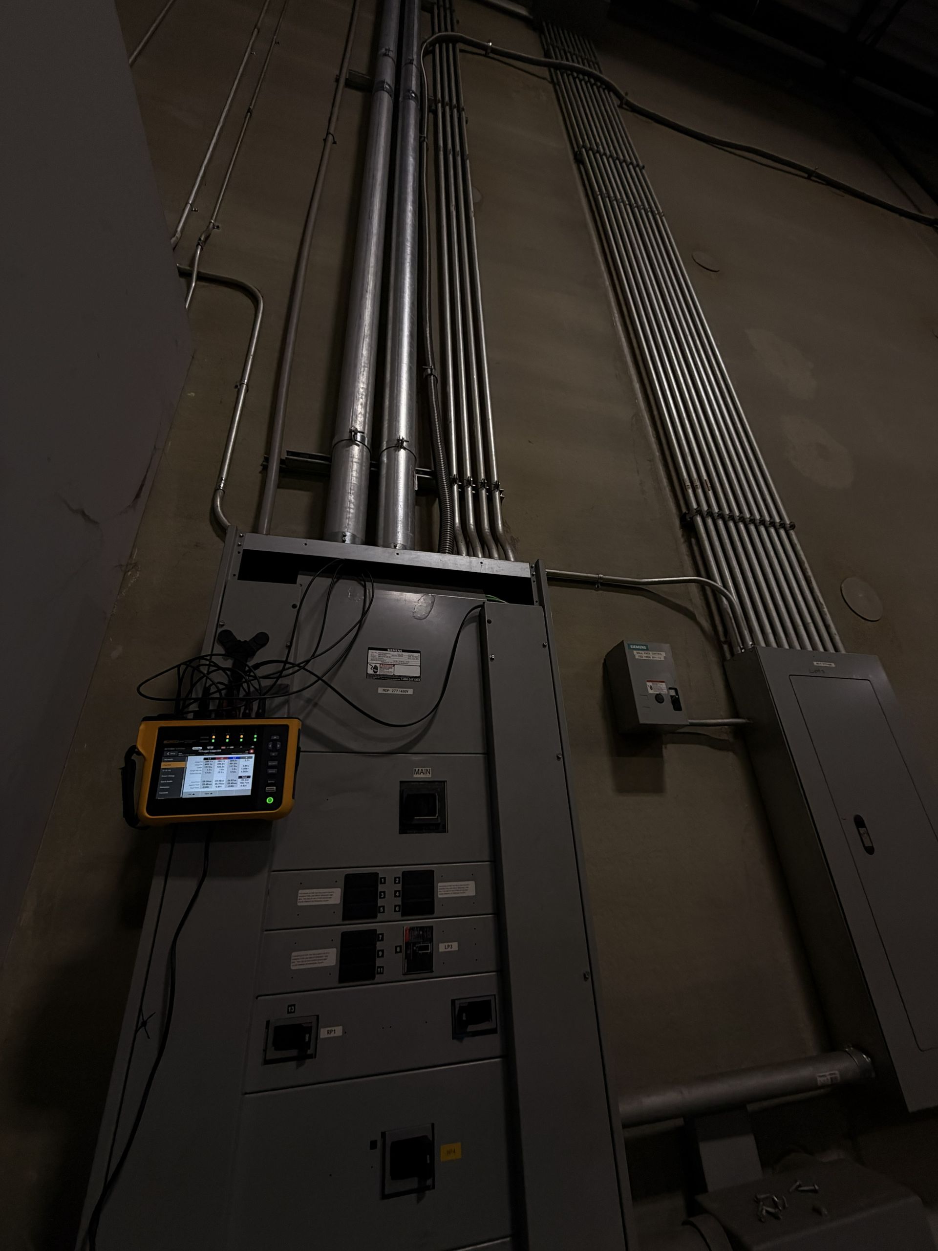 Electrical panel with pipes and conduit on a wall in a dim utility room