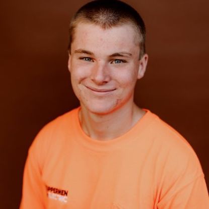 A person with short hair wearing a bright orange shirt against a solid brown background, smiling slightly.