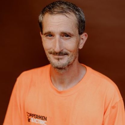 A person with short hair and a goatee, wearing an orange Coppermen Electric t-shirt against a brown background.