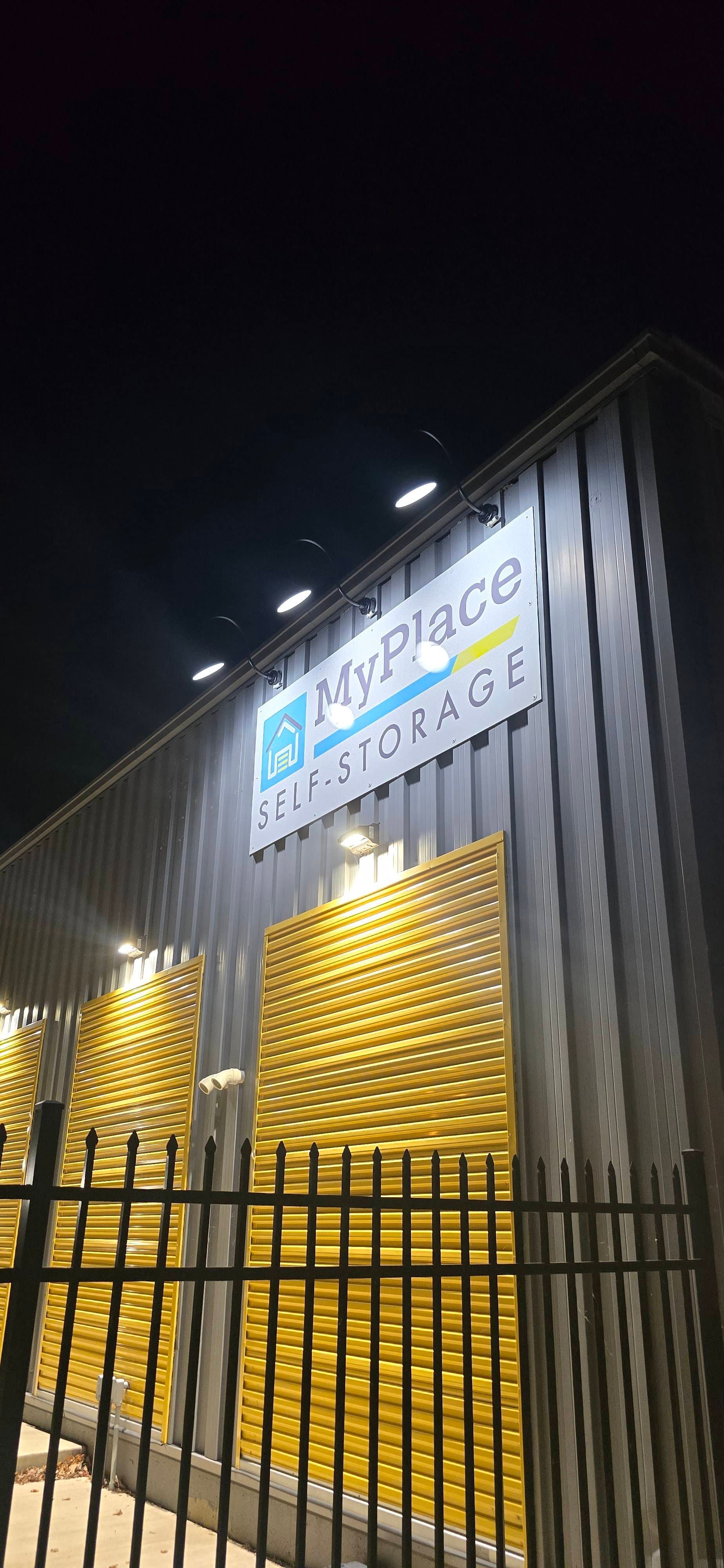 Nighttime view of a lit metal building facade with a sign above a yellow gate and fence.