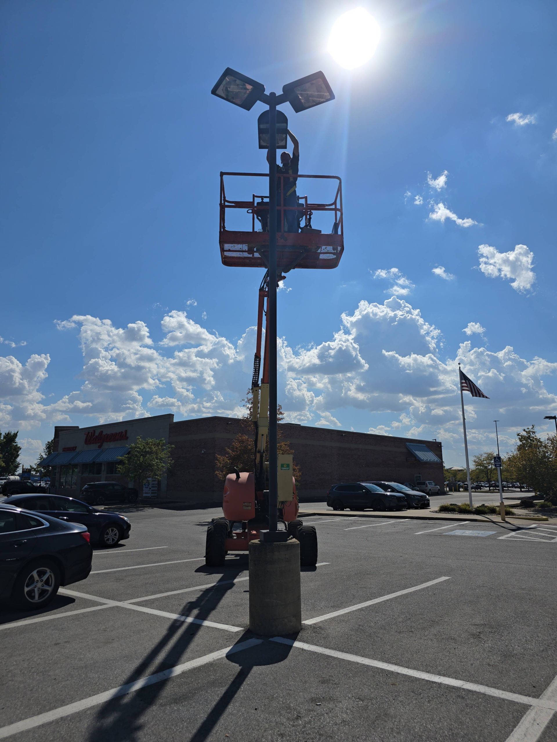 Parking lot light pole with solar panels and a lift basket under bright sun, with cars and a building behind