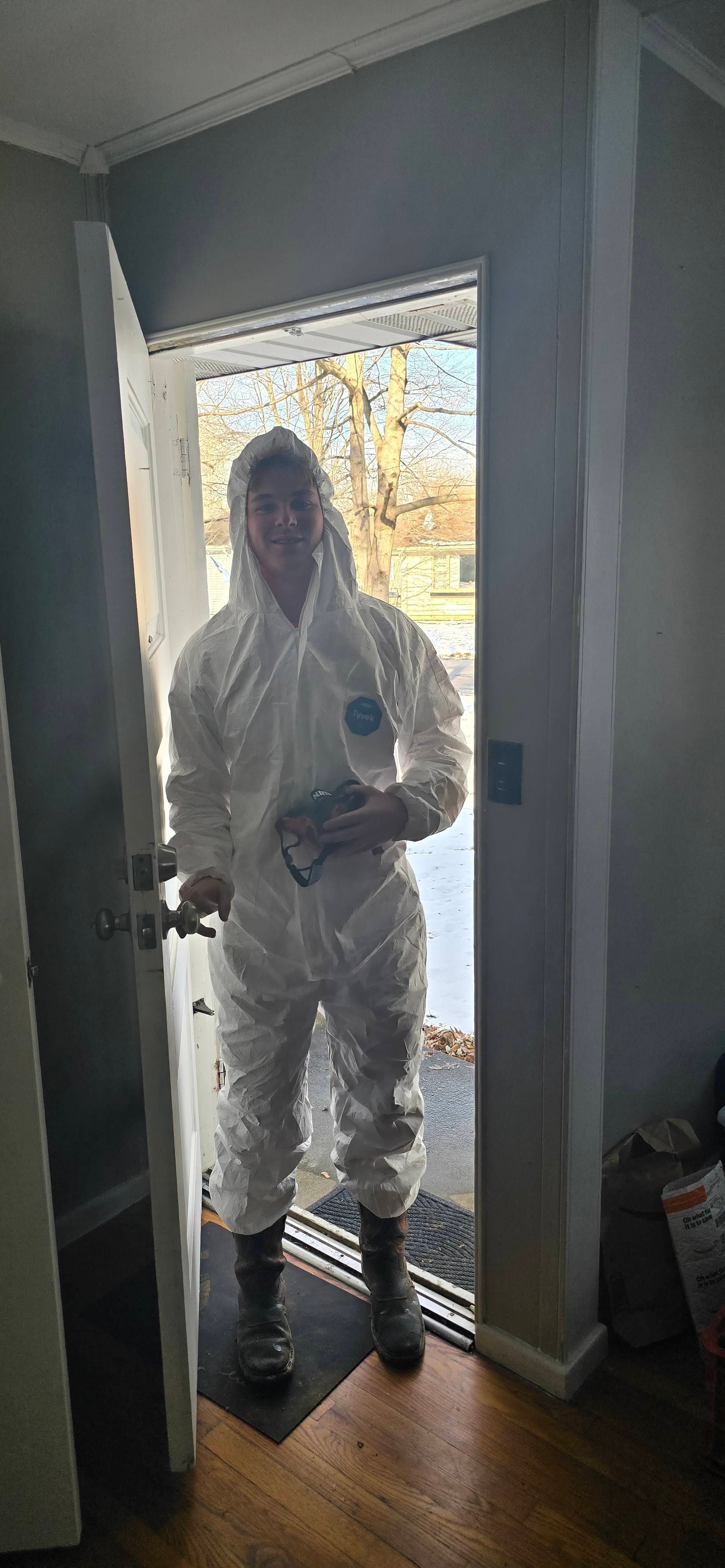 Person in white protective coveralls and hood standing in a doorway, holding a container and a tool.