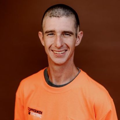 A person wearing a bright orange shirt with a company logo, smiling against a solid brown background.