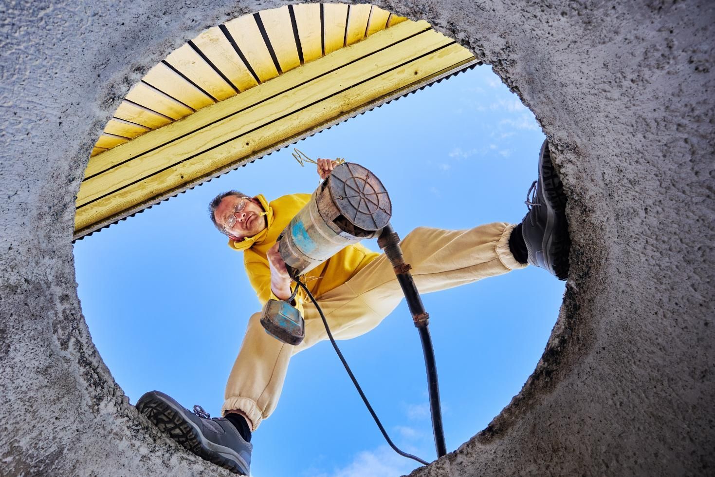Person standing over a circular opening, holding ropes against a blue sky and yellow structure overhead