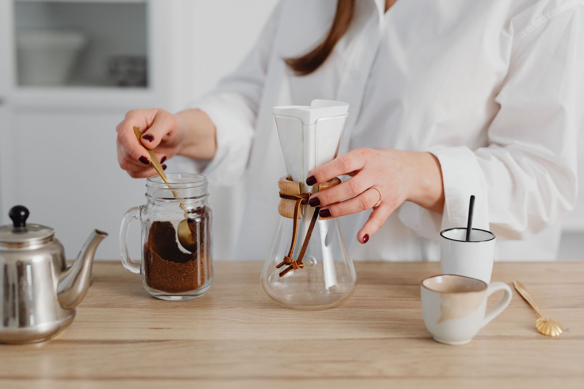 A woman standing behind a wooden counter scooping coffee grounds from a jar near a glass pour-over