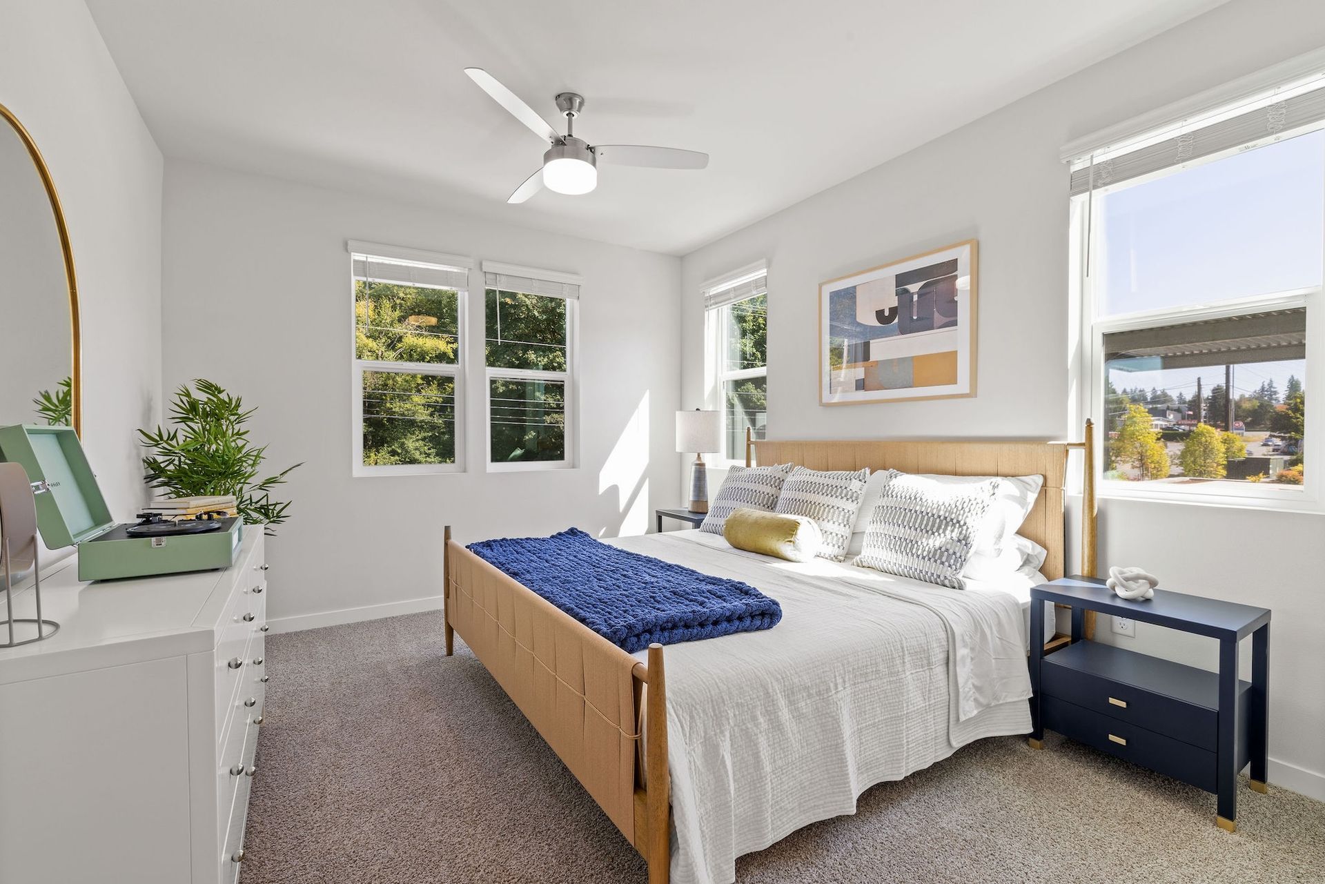 Bright bedroom with white walls, gray carpet, wooden bed, blue blanket, and two windows letting in daylight.