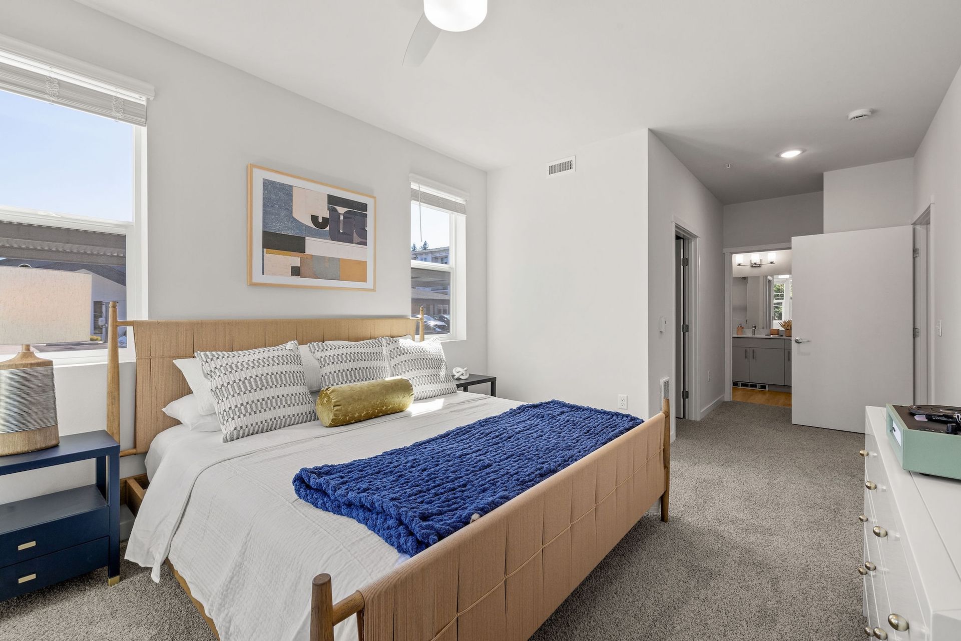 Bright bedroom with bed, blue throw, nightstands, wall art, and open doorway to hallway.