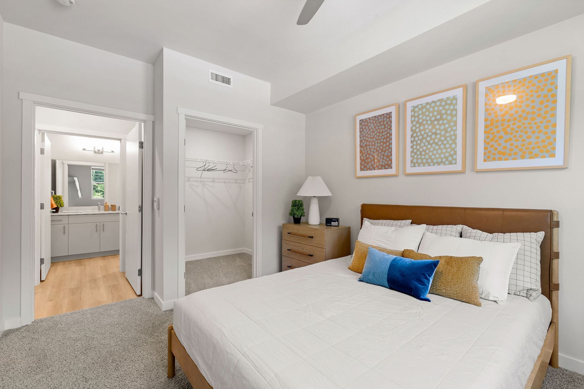 Bright bedroom with a bed, dresser, bedside lamp, and framed wall art; bathroom visible through doorway