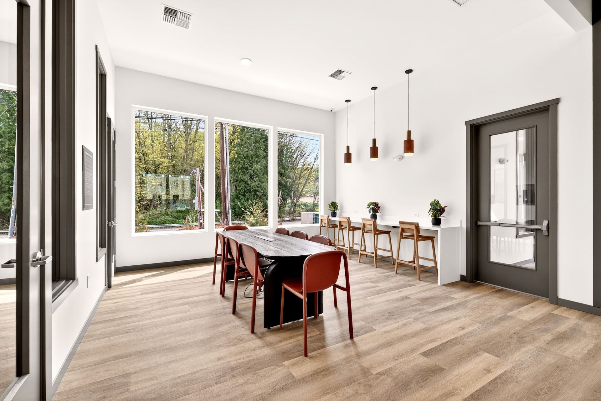Bright modern dining room with a long table, chairs, wood floors, and large windows overlooking trees