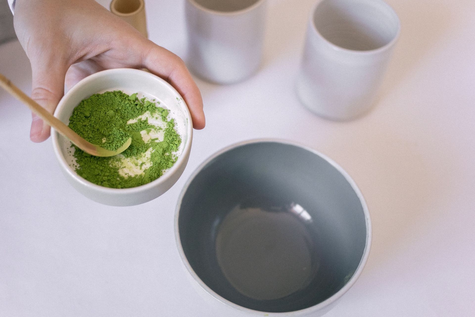 A hand holding a small bowl of green matcha powder with a spoon over a large grey bowl on a white surface.