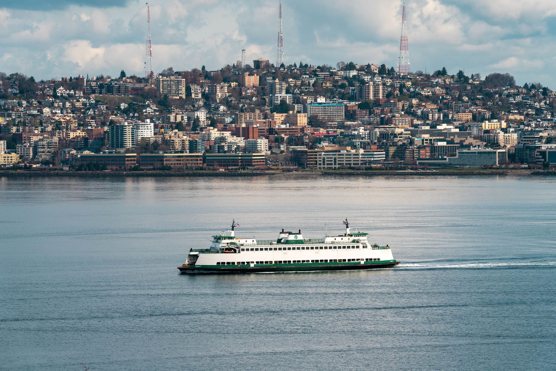 A white passenger ferry sailing on calm water with a dense city skyline on a hillside under a cloudy sky.