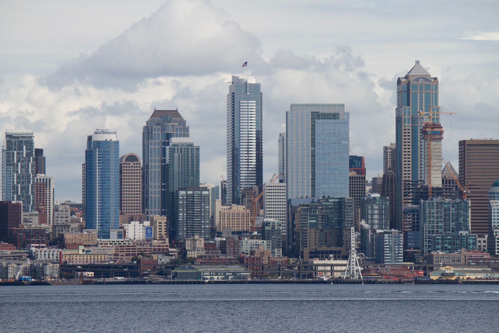 A view across water showing a dense city skyline filled with skyscrapers and high-rise buildings under cloudy skies.
