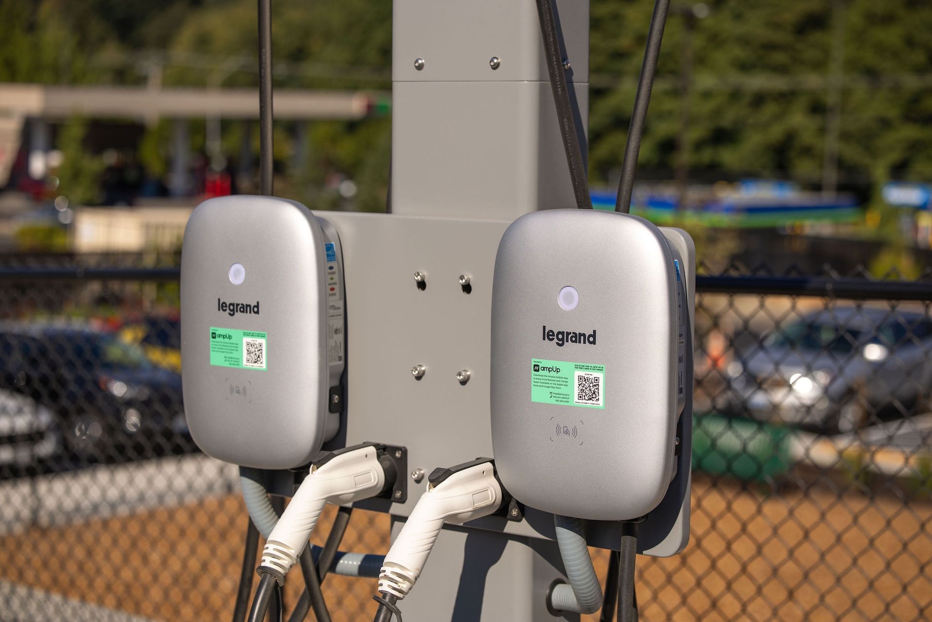 Two gray EV charging stations mounted on a pole beside a parking lot and chain-link fence.