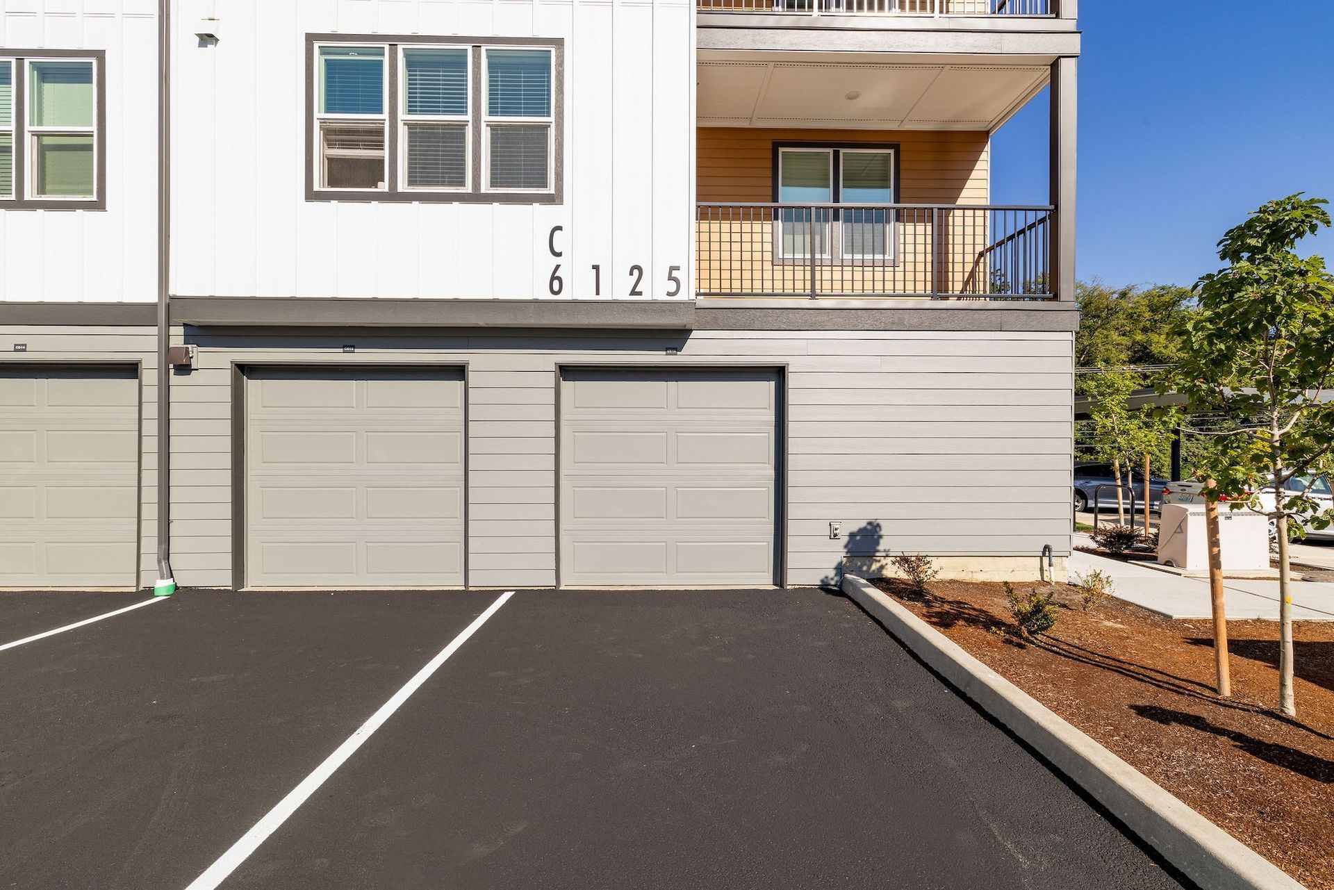 Apartment garage with three gray doors, black driveway, and balcony above in a sunny residential complex