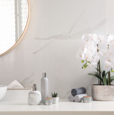 A bright bathroom vanity with a marble-patterned soap dispenser, lotion, small plant, candles, and stacked towels.